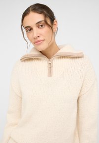 Sweter w kolorze kremowym, wykonany na drutach, z kołnierzem na pół zamek, posiadający kontrastowy beżowy zamek błyskawiczny i teksturę ściągacza. Podkreślony luźnym krojem.