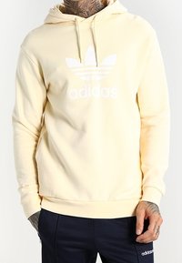 Žlutá mikina z měkkého materiálu, se bílým logem Adidas a třemi pruhy přes hruď. Kapuce se stahovací šňůrkou a žebrované manžety.