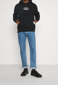 Svart hoodie med dragsko, ficka fram och vit logotyp; kombinerat med blå denimjeans, uppvikta benslut och svarta snörskor.