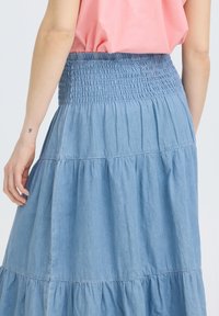 Frau trägt einen gestuften hellblauen Denimrock mit gerafftem Bund und ein in den Rock gestecktes pinkfarbenes ärmelloses Oberteil, von hinten gesehen.