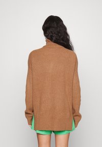 Brun ribbad stickad tröja med hög krage, med lös passform och sidoflikar, bärs över gröna shorts. Bakifrån.