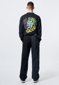 Camiseta negra de manga larga con gráficos de texto coloridos en la parte posterior. Combinada con pantalones de mezclilla negros holgados y zapatos de color claro.