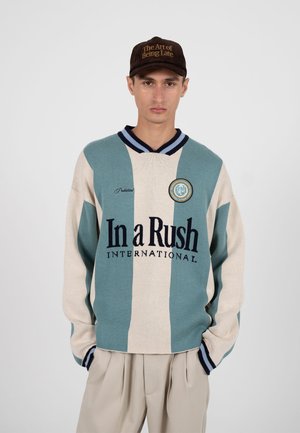 Homme portant un pull à rayures beige et bleu sarcelle avec le texte "In a Rush International", un pantalon beige et une casquette noire arborant l'inscription "The Art of Being Late".