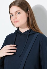 Giovane donna con capelli castani chiari e lisci che indossa una felpa con cappuccio blu scuro, mentre tocca il lacciolo della felpa, su uno sfondo bianco neutro.