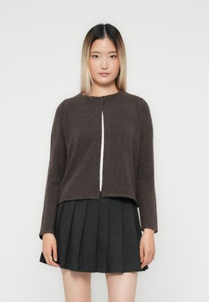 Jonge vrouw met steil ombre haar, gekleed in een donkerbruine lange mouwen cardigan met knopen en een zwarte geplooide rok, tegen een witte achtergrond.