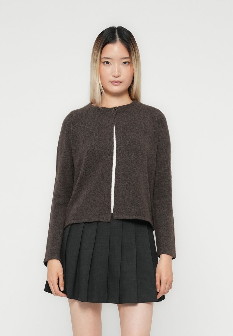Giovane donna con capelli lisci ombre che indossa un cardigan lungo a maniche, button down, marrone scuro e una gonna nera plissettata, su sfondo bianco.