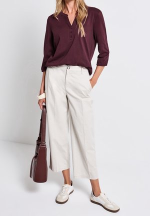 Top henley de manga larga en color burdeos con botones en la parte delantera, combinado con pantalones anchos de pierna en beige claro y zapatillas blancas, complementado con un bolso marrón.