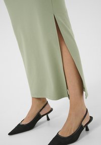 Robe verte claire, longueur genou avec fentes sur les côtés; associée à des chaussures slingback noires à bout pointu avec un tissu texturé et un petit talon.