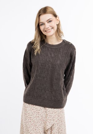 Młoda kobieta o blond włosach uśmiechająca się, ubrana w brązowy rozpinany sweter z guzikami na ramieniu oraz jasną spódnicę w kwiaty.