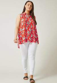 Rote ärmellose Bluse mit weißem Blumenmuster, Kragen und asymmetrischem Saum; kombiniert mit weißen Slim-Fit-Hosen und schwarzen Ballerinas.
