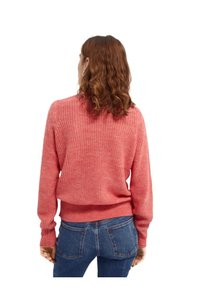 Suéter de punto en color coral con corte holgado, puños y bajo acanalados. La vista trasera muestra un patrón texturizado y una tela suave contra unos jeans azules.