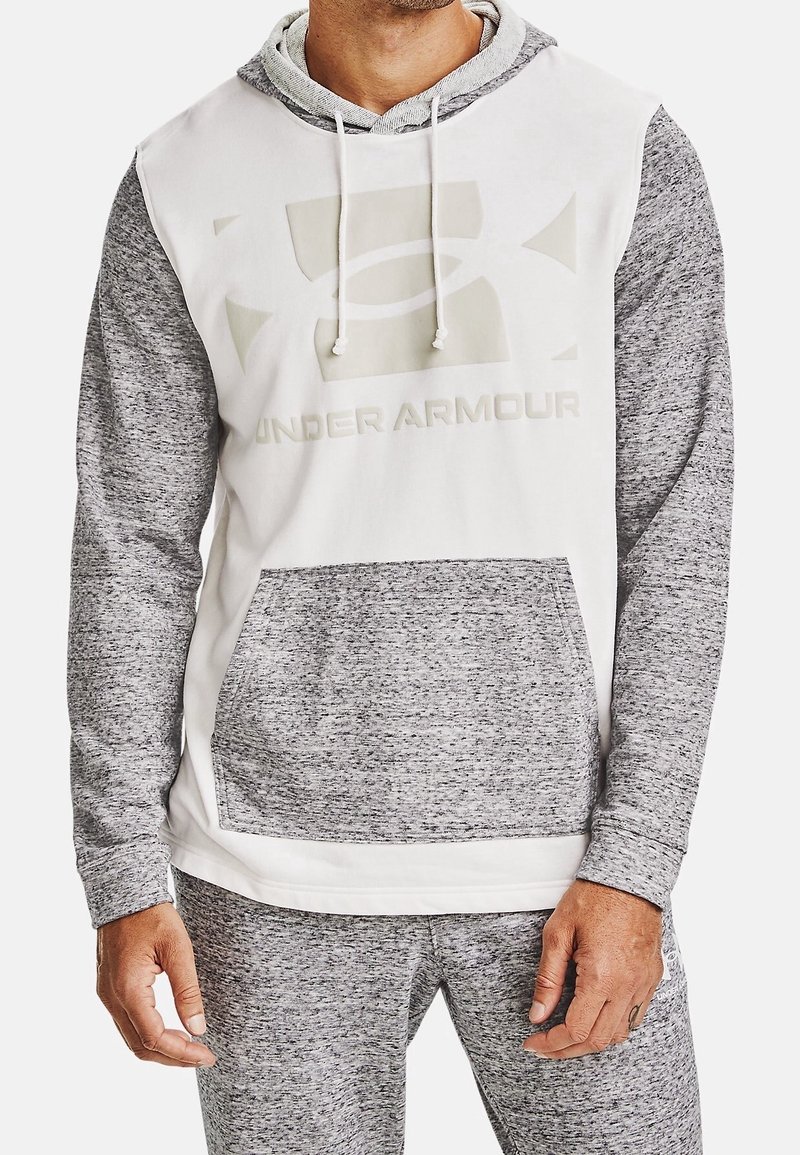 Homme portant un sweat à capuche bicolore gris et blanc avec un logo frontal et une poche kangourou apparente, accompagné d'un pantalon de survêtement assorti.