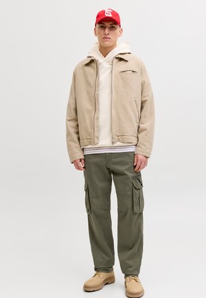 Jonge man met een rode pet, beige jas, crème hoodie, olijfgroene cargobroek en tankleurige laarzen, staand tegen een effen lichte achtergrond.