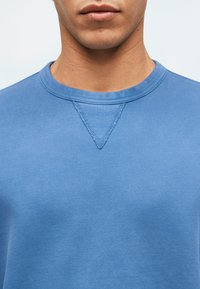 Κοντινό πλάνο του κάτω μέρους του προσώπου και του λαιμού ενός ατόμου που φοράει μπλε φούτερ με στρογγυλή λαιμόκοψη και ραμμένο τριγωνικό σχέδιο κάτω από τον γιακά.