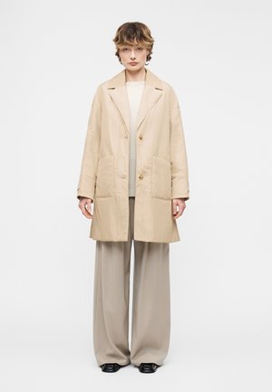 Person iført en beige oversized frakke med store forlommer, brede beige bukser, hvid top og sorte sko, stående mod en ensfarvet baggrund.