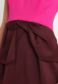 Gros plan d'une robe avec un haut ajusté rose vif et une jupe bordeaux foncé avec un grand nœud plié à la taille.