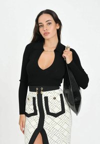 Donna con capelli lunghi e scuri che indossa una maglia nera a maniche lunghe con scollo profondo, gonna bianca con motivo e spacco frontale, che tiene una borsa nera con texture.