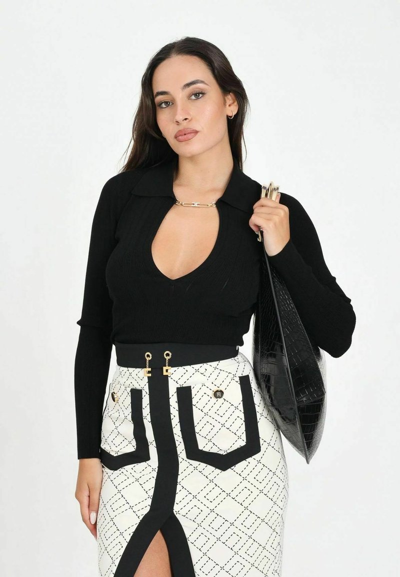 Donna con capelli lunghi e scuri che indossa una maglia nera a maniche lunghe con scollo profondo, gonna bianca con motivo e spacco frontale, che tiene una borsa nera con texture.