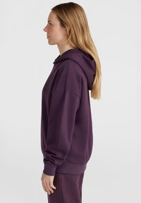 Felpa viola con una grande tasca frontale, cappuccio con cordoncino e polsini a costine. Indossata con pantaloni della tuta coordinati, con un logo sulla gamba.