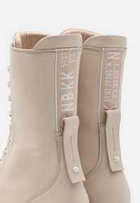 Paire de bottines en cuir beige à lacets, avec des languettes en tissu ornées de texte blanc brodé et des détails de rivets.