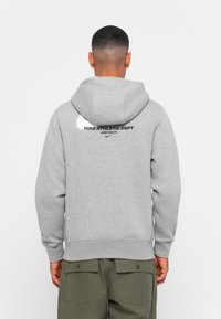 Grijze hoodie met een trekkoord capuchon, zachte textuur. Heeft op de achterkant de tekst "NIKE ATHLETIC DEPT" en "JUST DO IT" in het zwart gedrukt.
