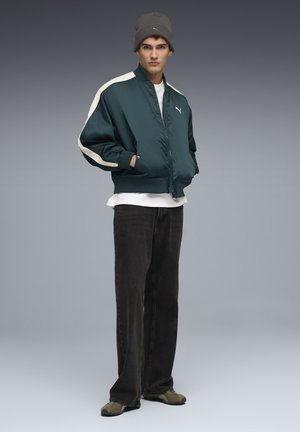 Teal bomberjakke med lysebeige detaljer, parret med en hvid t-shirt, sorte jeans og olivengrønne sko. Modellen bærer en grå beanie.