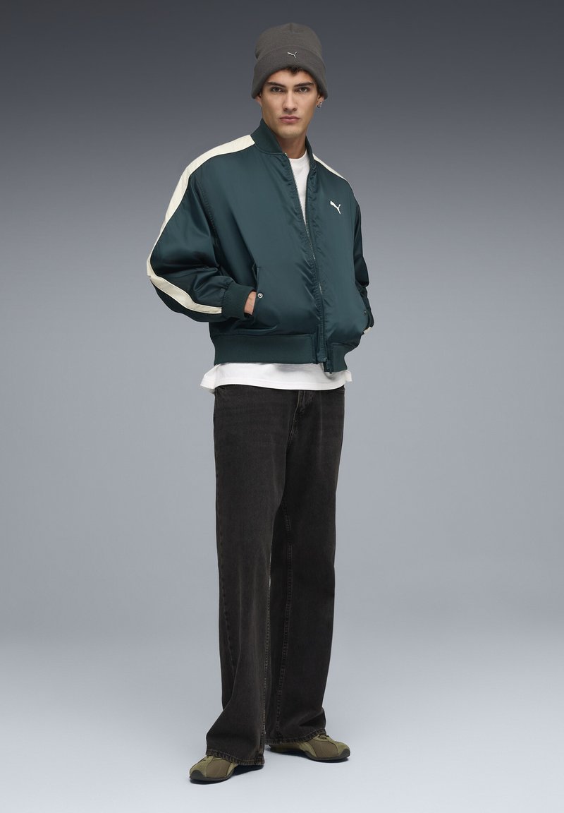 Teal bomberjack met lichtbeige accenten, gecombineerd met een wit T-shirt, zwarte jeans en olijfgroene schoenen. Model draagt een grijze beanie.
