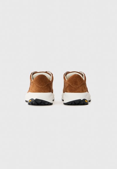 O pereche de teniși din suede maro cu tălpi intermediare albe și tălpi exterioare negre Vibram, văzuți din spate pe un fundal simplu și deschis.