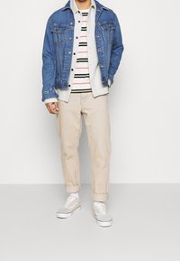 Denimjasje, grijze hoodie, gestreept shirt met oranje en groen, beige opgerolde broek en grijs-witte sneakers met crème accenten.