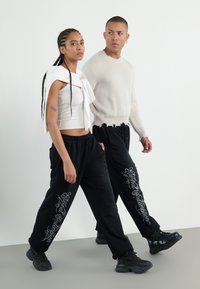 Black sweatpants with white graphic text, paired with a cropped white tank top and a cream sweater. Both models wear black sneakers.