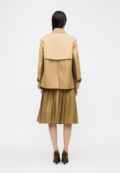 Γυναίκα με μακριά σκούρα μαλλιά φοράει καφέ κοντό μπουφάν, πλισέ midi φούστα και καφέ τακούνια, στέκεται με την πλάτη γυρισμένη σε απλό λευκό φόντο.