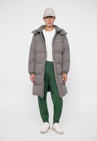 Abrigo de plumón gris con diseño acolchado, de largo hasta la cadera y con capucha, combinado con pantalones verdes y zapatillas blancas. La gorra añade un acento neutro.