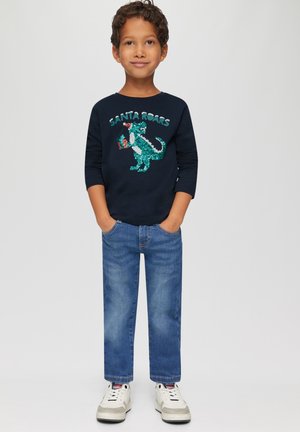 Marineblaues Langarmshirt mit einem glitzernden grünen Dinosaurier-Motiv, das "SANTA ROARS" sagt; kombiniert mit hellblauen Jeans und weißen Sneakers.