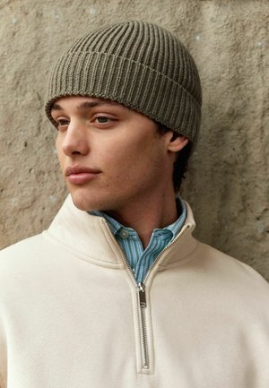 Olive green ribbed knit beanie, paired with a cream zip-neck sweater over a light blue striped shirt. Textured background adds depth.