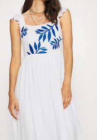 Vrouw met een witte mouwloze jurk met blauwe bladborduursels op de borst en meerdere gouden kettingen, gefotografeerd tegen een effen achtergrond.