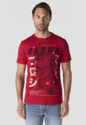 Hombre vistiendo una camiseta roja con diseño de dragón negro y texto que dice "Osaka" y caracteres japoneses, combinado con vaqueros azul claro.