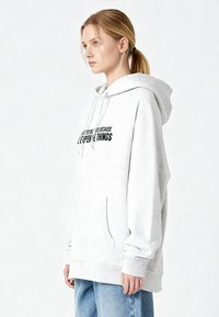 Felpa con cappuccio grigio chiaro con tasca a marsupio frontale, cappuccio con coulisse e testo nero stampato sul petto. Tessuto morbido con finitura spazzolata.