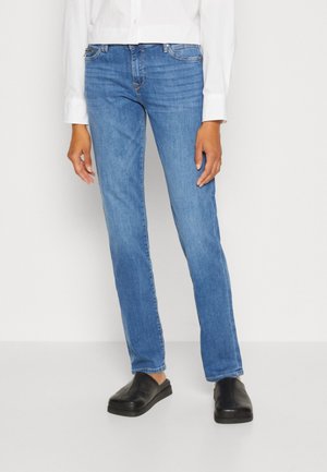 Personne portant un jean droit bleu, une chemise blanche à manches longues et des chaussures noires à enfiler, se tenant devant un fond clair uni.
