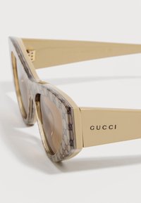 Gafas de sol beige de Gucci con lentes tintadas marrones y montura gruesa con patrón que presenta el logo de Gucci en la patilla.