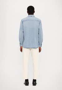 Camisa vaquera azul claro con cuello puntiagudo, puños abotonados y dobladillo curvado. Se combina con pantalones de color crema y zapatos negros.