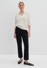 Hellebeige V-Ausschnitt-Strickjacke mit kurzen Ärmeln, kombiniert mit schwarzer, taillierter Hose. Das Outfit besticht durch glatte Texturen und eine entspannte Passform. Schwarze Ballerinas.