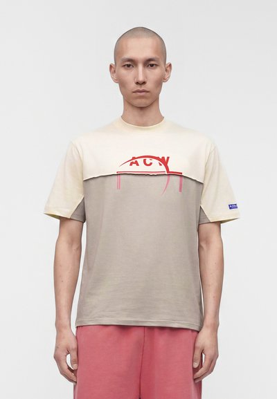 Homme à la tête rasée portant un t-shirt beige et crème en bloc de couleurs avec un motif rouge, short rose, debout devant un fond blanc uni.