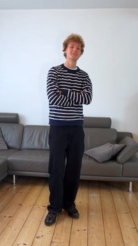 Randig marinblå och vit tröja, svarta byxor och mörka skor. Står avslappnat med armarna i kors i ett minimalistiskt rum med trägolv.