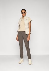 Lichtbeige gewatteerd vest over een beige shirt, gecombineerd met donkerbruine broek en beige sneakers, gedragen met een zonnebril. Eenvoudig, functioneel ontwerp.