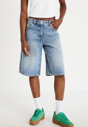 Person iført knælange lyseblå denimshorts, hvide sokker, grønne sneakers og en hvid cropped top, stående med den ene hånd i lommen.