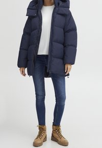 Námořnická puffer bunda s kapucí, zipovým uzávěrem a náprsními kapsami. Nositá přes bílý top, v kombinaci s modrými džíny a hnědými šněrovacími botami.