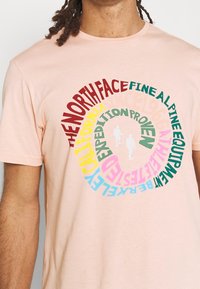 Světle růžové bavlněné tričko s kulatou multicolor textovou grafikou. Text zahrnuje název značky a fráze, s dvěma postavami v bílé siluetě.