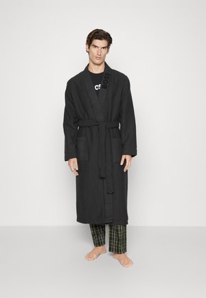 Zwarte, gestructureerde badjas met een strikceintuur, zijzakken en een subtiele "BOSS" branding op de kraag, gedragen over een geruite broek.