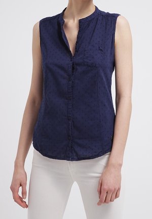 Mouwloze marineblauwe blouse met knopen, gerimpelde kraag en subtiel gestippeld patroon, gedragen met een witte broek.