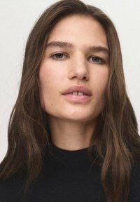 Close-upportret van een persoon met mediumbruin, golvend haar, gekleed in een zwarte turtleneck. Kenmerken zijn een lichte huid en natuurlijke make-up.